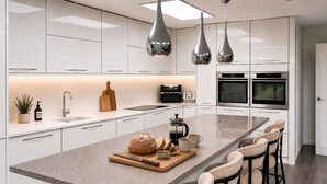 A modern kitchen with white cabinetry, a central island with bar stools, a coffee maker, and a bread loaf. - Modern coastal retreat – 5 bed home offering space and style for up to 12 guests (Folkestone)