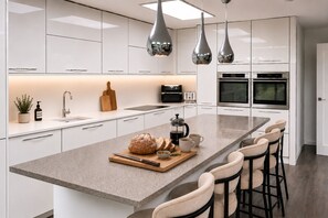 A modern kitchen with white cabinetry, a central island with bar stools, a coffee maker, and a bread loaf. - Modern coastal retreat – 5 bed home offering space and style for up to 12 guests (Folkestone)