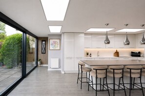A modern kitchen with a central island, pendant lighting, and a view through large windows. - Modern coastal retreat – 5 bed home offering space and style for up to 12 guests (Folkestone)