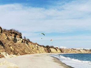 Uma praia com falésias, uma faixa de areia e vista para edifícios ao longe.