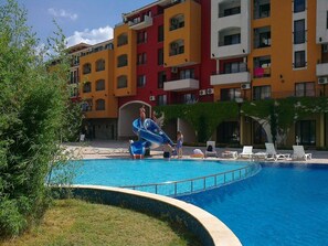 En swimmingpool med vandrutsjebane, omgivet af et hotel med flere balkoner og en fliseterrasse.
