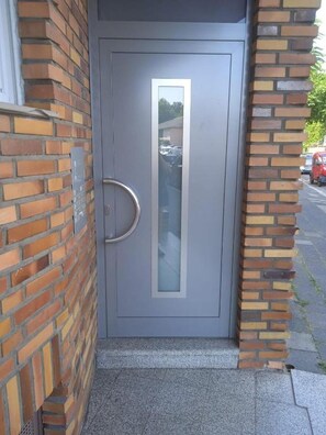 A grey door with a vertical handle and a glass panel, set in a brick wall with a metal plate on the left. - Kaiserslautern City Center Apartment · 3 Rooms · Sleeps 5 · Full Kitchen (Kaiserslautern)