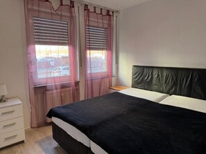 Une chambre à coucher comprenant un lit, une table de chevet, une commode et une fenêtre avec des stores.