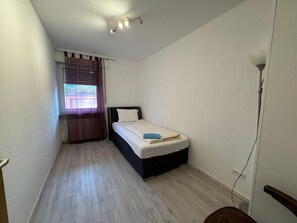 Une petite chambre avec un lit simple, un plancher en bois, une fenêtre avec des rideaux et un luminaire fixé au mur.