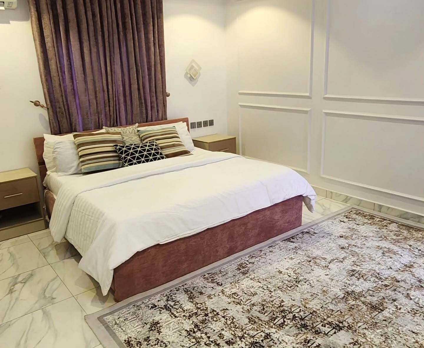 A neatly made bed with a white comforter and multiple pillows, a wooden nightstand with a drawer, a patterned rug on the floor, and a wall-mounted light fixture.