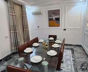 A dining room with a glass table, brown leather chairs, white plates, and a painting on the wall. - Nomads Nest Retreat (Abuja)