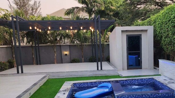 Une piscine avec un jacuzzi, une pergola illuminée de guirlandes lumineuses et un petit espace clos avec une porte.