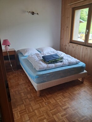Une chambre avec un plancher en bois, un lit avec un matelas bleu, des oreillers blancs et une lampe rose posée sur une table de chevet en bois.