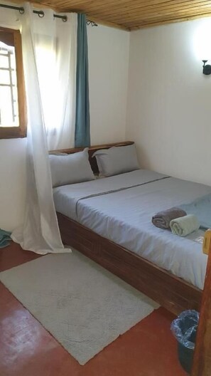 A single bed with a wooden frame, a white bedspread, and a blue curtain partially drawn. - Chez Rasoa Andasibe (Andasibe)