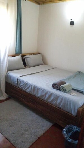 A single bed with a wooden frame, a white mattress, and a blue blanket. There is a small trash bin to the right of the bed. - Chez Rasoa Andasibe (Andasibe)