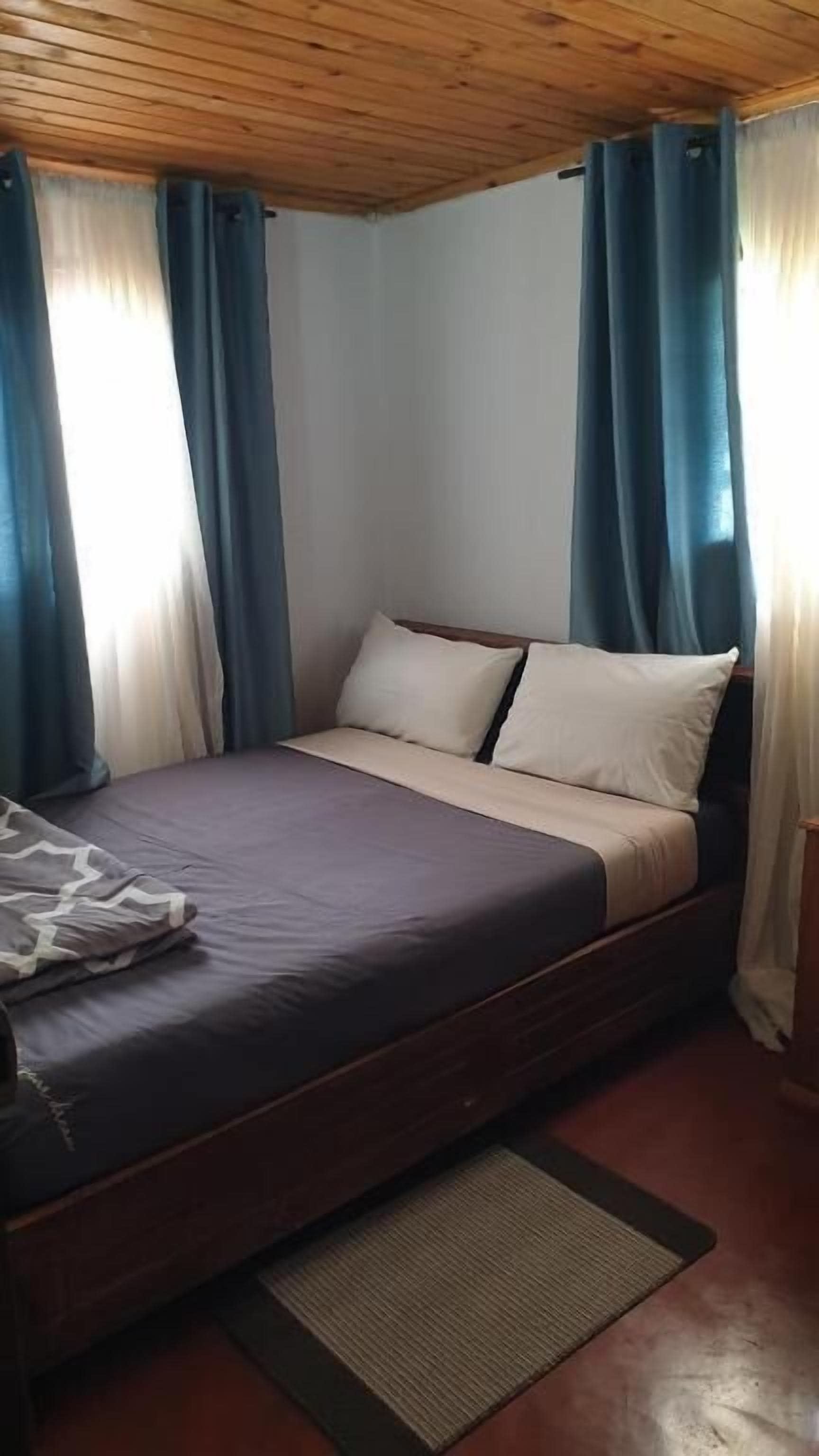 Un lit simple avec une tête de lit en bois, un matelas de couleur foncée et des oreillers blancs. Des rideaux bleus sont accrochés aux fenêtres, et on aperçoit un petit tapis sur le sol.