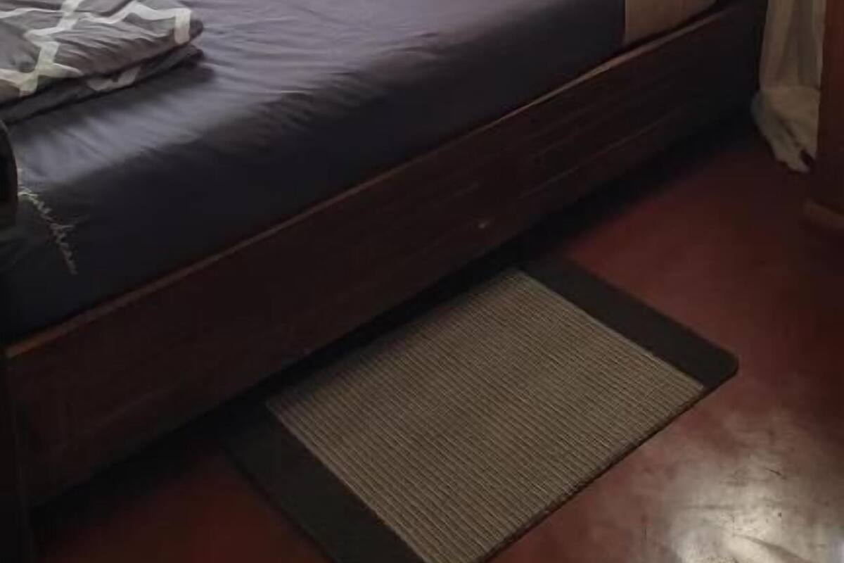 Ein Einzelbett mit Holz-Kopfteil, einer dunkelgrauen Matratze und weißen Kissen. Blaue Vorhänge sind an den Fenstern, und ein kleiner Teppich ist auf dem Boden zu sehen.
