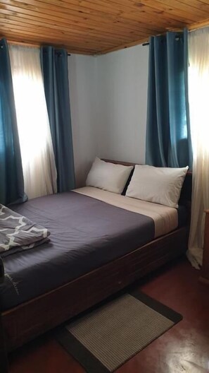 Un lit simple avec une tête de lit en bois, un matelas de couleur foncée et des oreillers blancs. Des rideaux bleus sont accrochés aux fenêtres, et on aperçoit un petit tapis sur le sol.