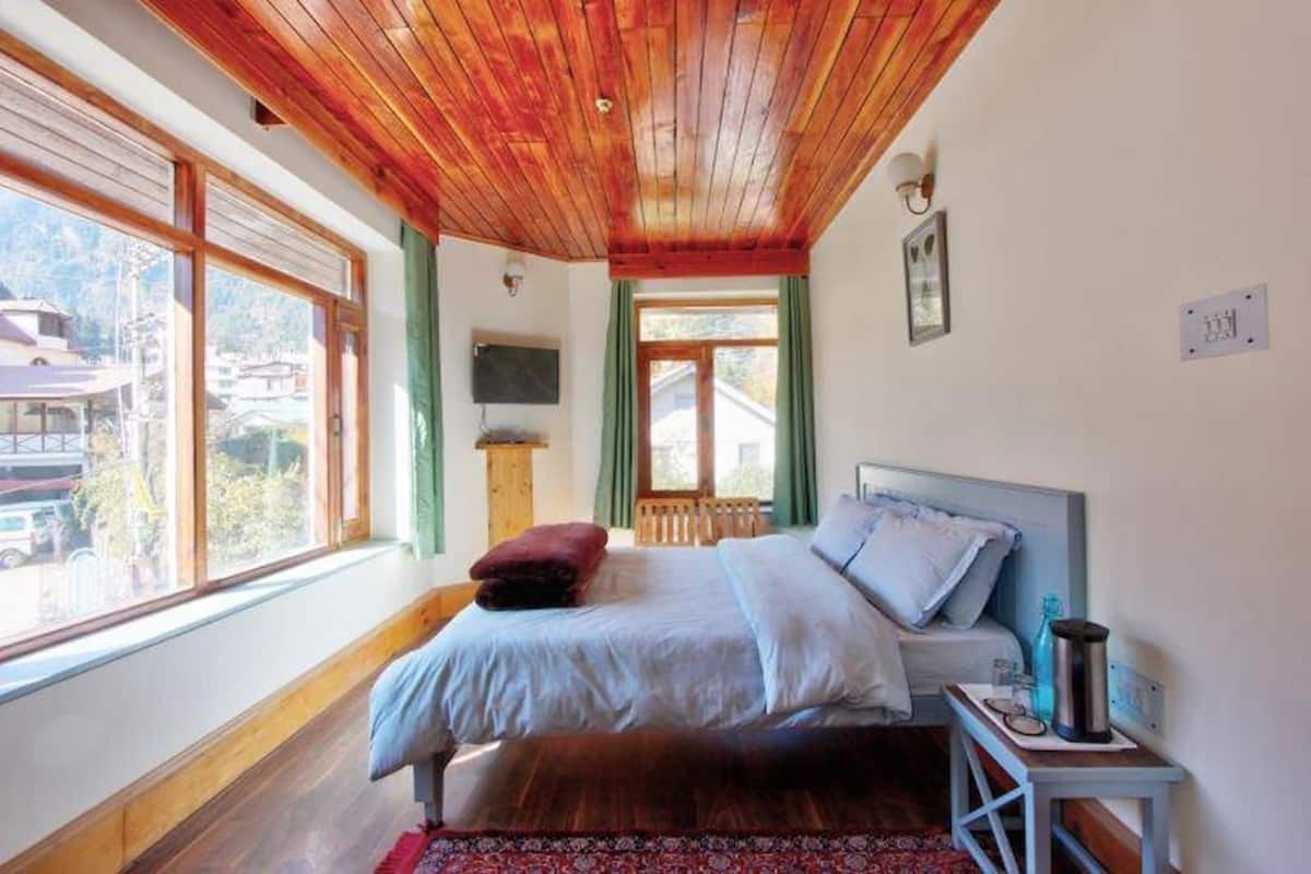 A bedroom with a wooden ceiling, a bed with pillows, a bedside table with a water bottle, and a window with a view of buildings.