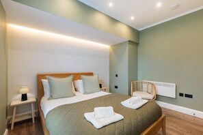 A neatly arranged hotel room with a bed, bedside tables, a chair, and a wall-mounted heater. - Luxurious & spacious central modern 2bed flat (London)