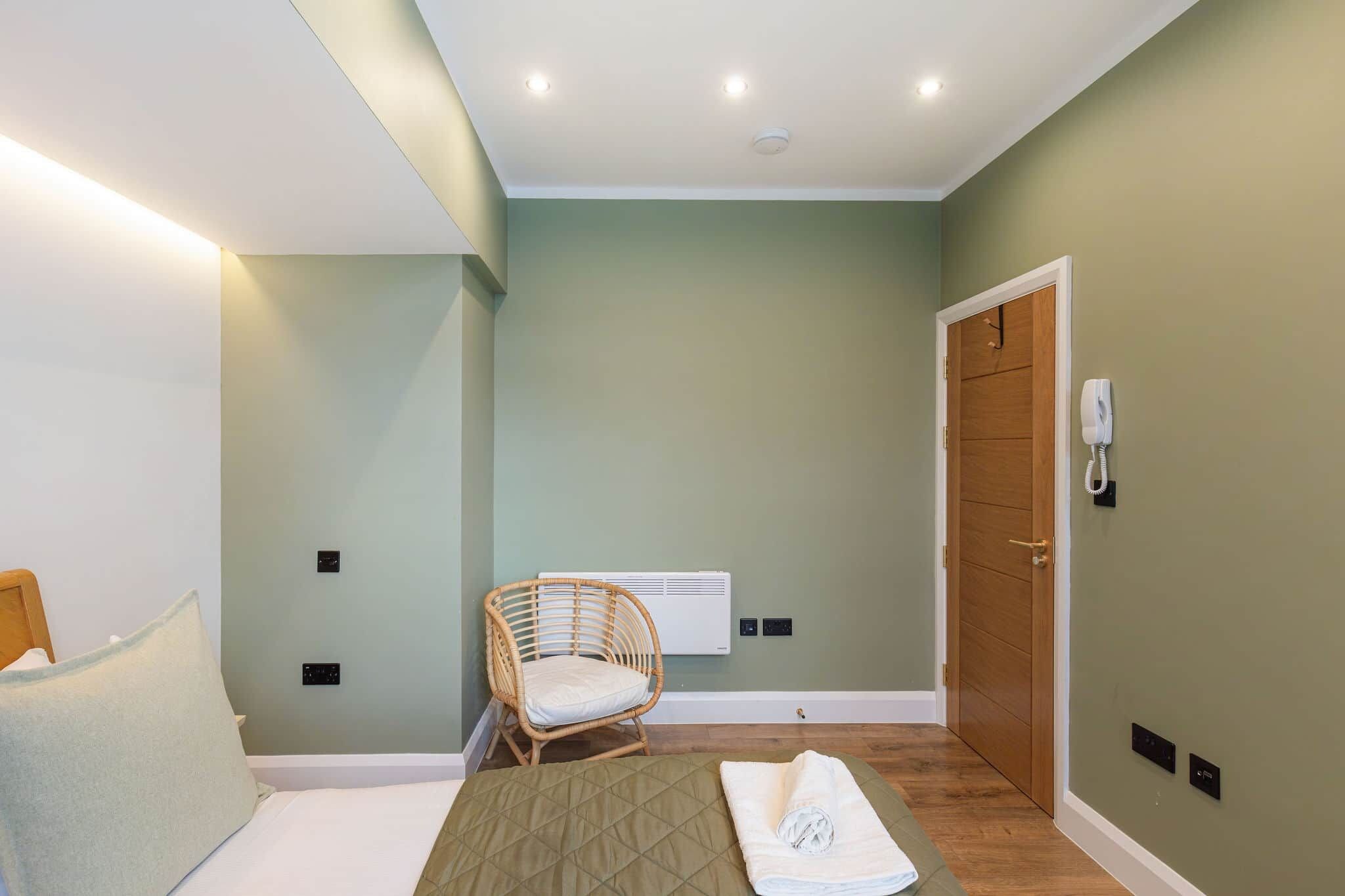 A bedroom with a bed, a wicker chair, a wooden door, and a wall-mounted phone.