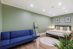 A bedroom with a bed, a blue sofa, a bedside table, a lamp, and framed artwork on the wall. - Stunning spacious modern central flat free parking (London)
