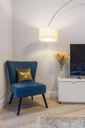 A blue chair with a bee pillow, a white side table with a vase, and a modern floor lamp. - Stylish 2BR Georgian Flat • Prime Camden Location (London)