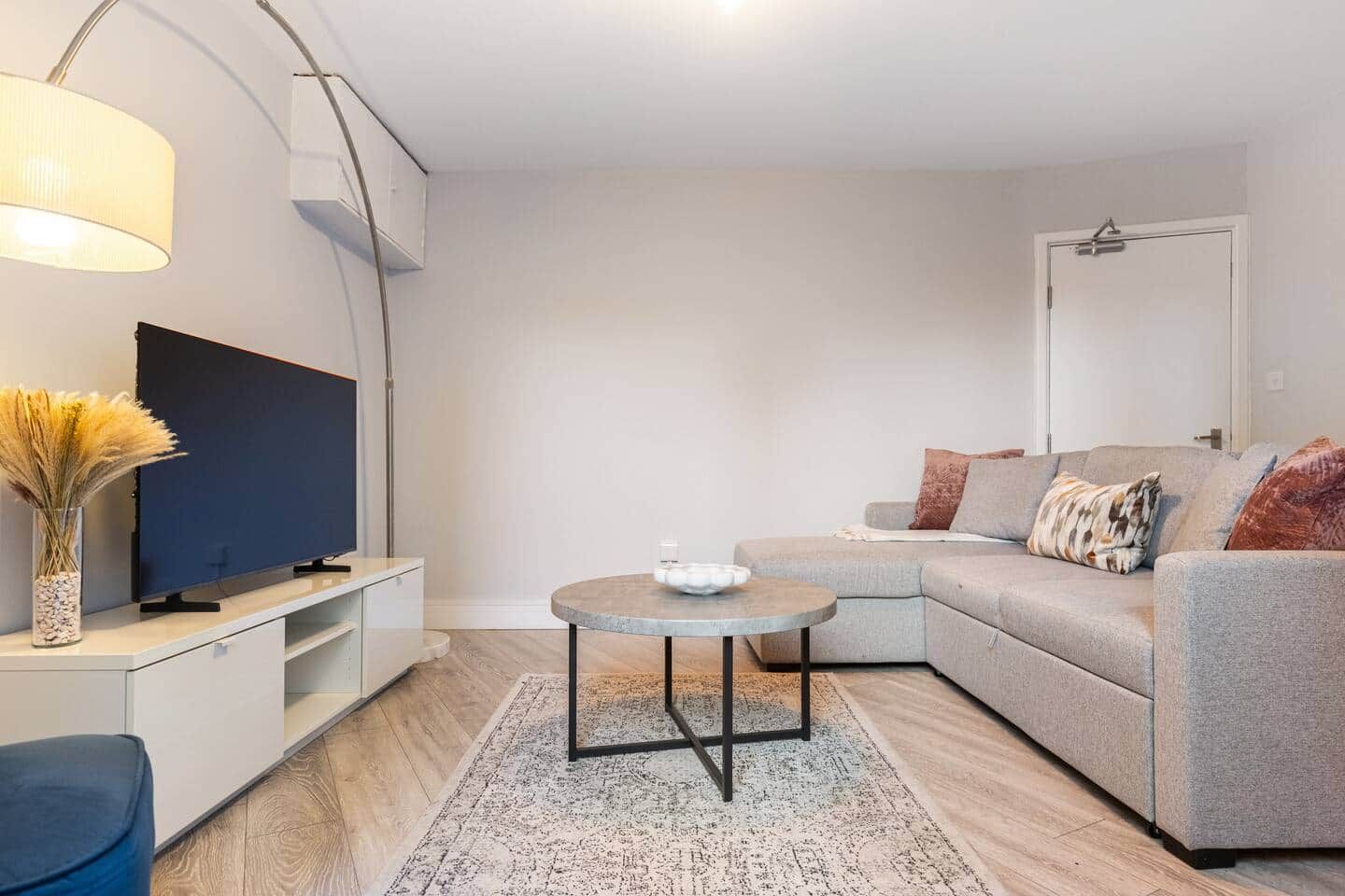 A modern living room with a flat-screen TV, a round coffee table, a grey sofa, and a decorative vase.
