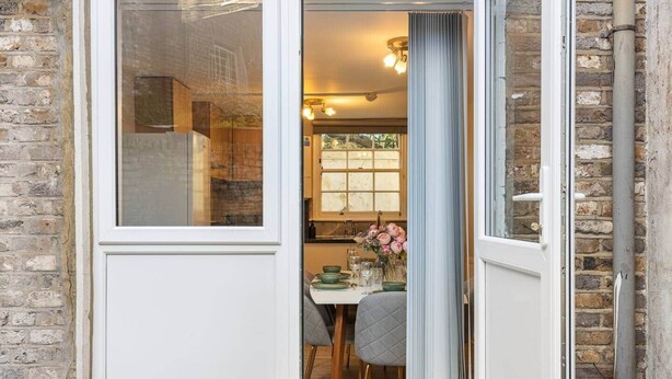Une porte qui mène à une salle à manger où une table est dressée pour deux personnes, une fenêtre offrant une vue agréable et un mur en briques.