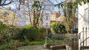 Un jardin avec un chemin pavé menant à l’entrée d’un bâtiment, entouré de verdure et délimité par une clôture en métal.