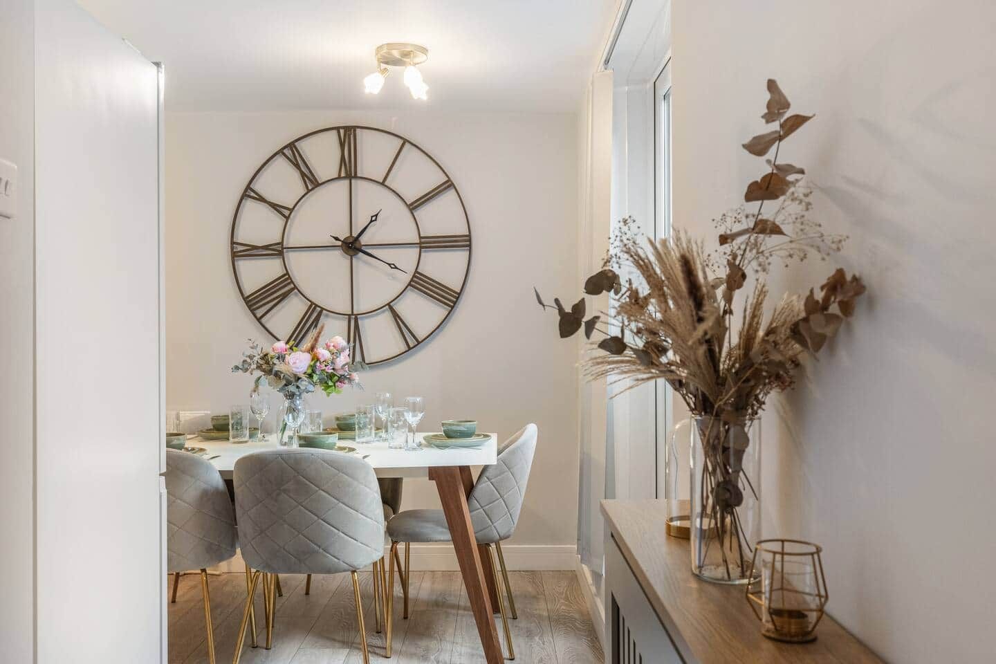 Un comedor con un reloj de pared grande, una mesa dispuesta para dos personas y un jarrón con flores secas.