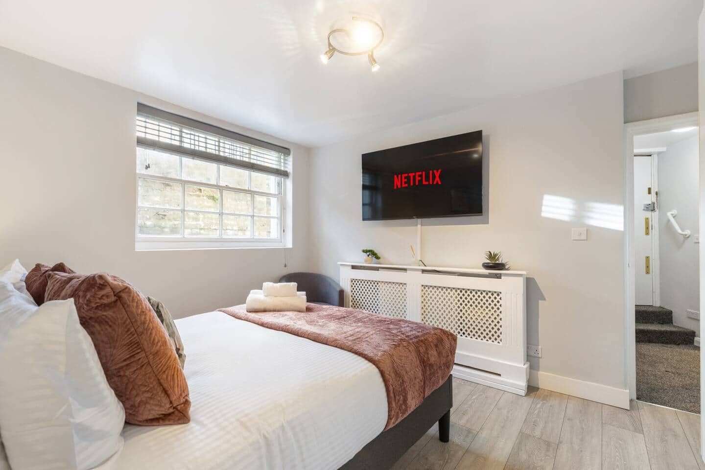 A bedroom with a bed, a TV mounted on the wall, a window, and a door leading to a staircase.