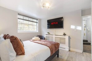 A bedroom with a bed, a TV mounted on the wall, a window, and a door leading to a staircase. - Stylish 2BR Georgian Flat • Prime Camden Location (London)