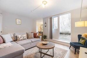 A modern living room with a grey sofa, a round coffee table, and a view of the outdoors through a large window. - Stylish 2BR Georgian Flat • Prime Camden Location (London)