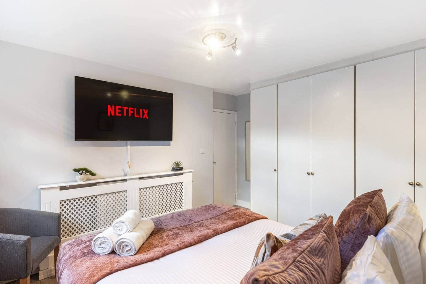 A hotel room with a bed, a TV mounted on the wall, a wardrobe, and a seating area with a chair.