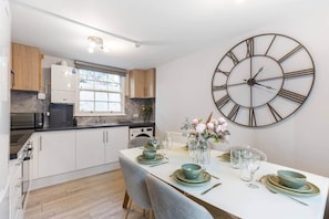 A modern kitchen with white cabinets, a large wall clock, and a dining area set for four with a vase of flowers. - Stylish 2BR Georgian Flat • Prime Camden Location (London)