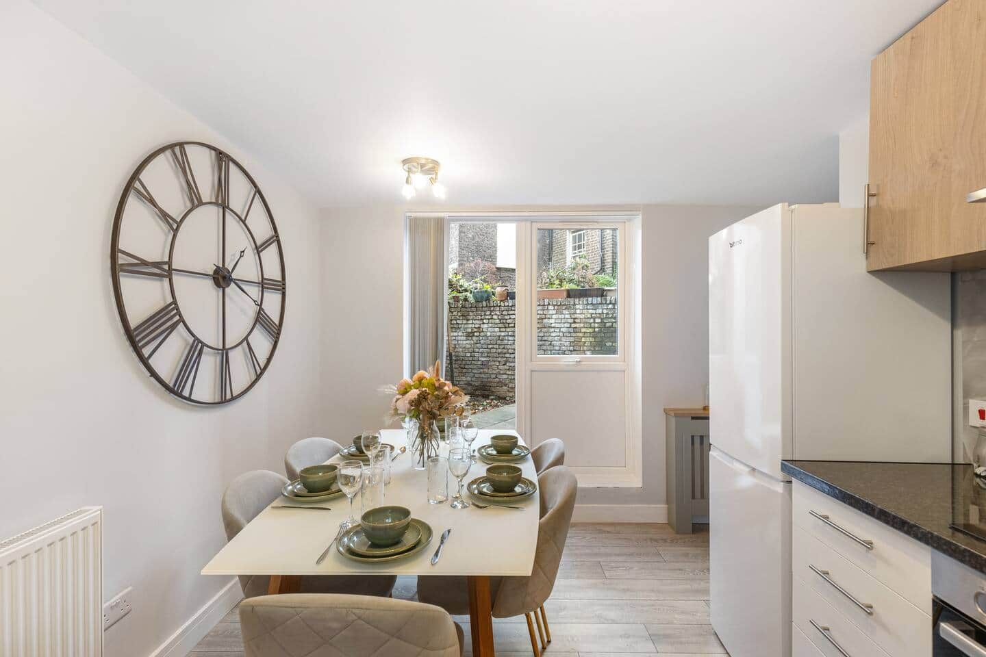 A compact kitchen with a dining area, a large wall clock, and a view through a window.