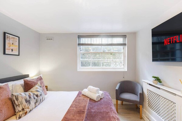 A bedroom with a bed, a chair, a window, a radiator, and a wall-mounted TV. - Stylish 2BR Georgian Flat • Prime Camden Location (London)