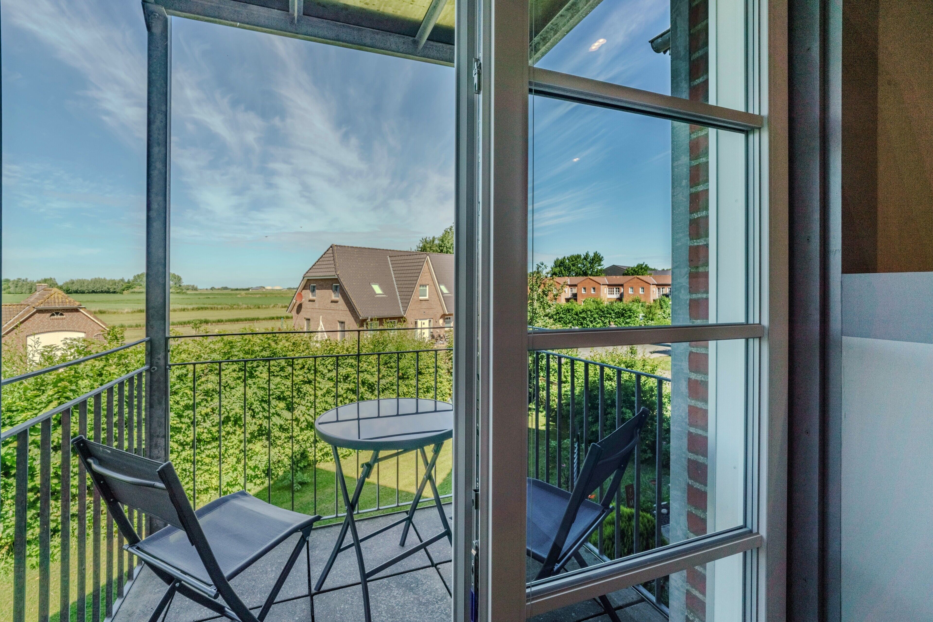 Ein Balkon mit Tisch und Stühlen, Blick auf Häuser und Grünflächen, und ein klarer Himmel.