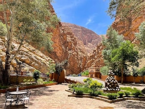 A courtyard with a stone table and chairs, surrounded by greenery and a mountain backdrop.