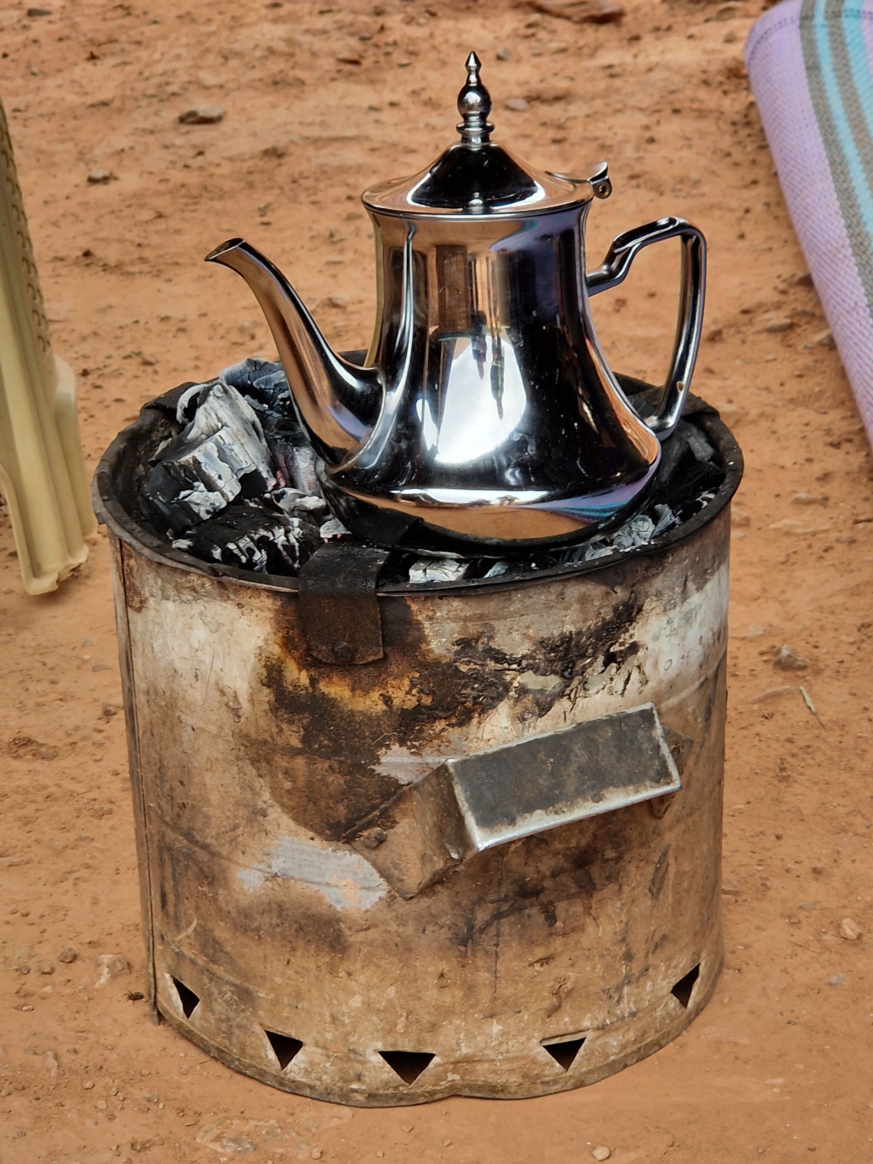 A portable stove with a metal pot and a lid, placed on a dirt surface.