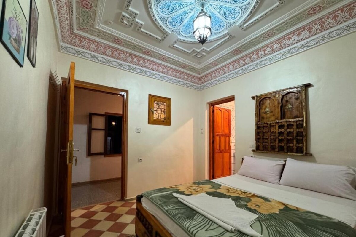 Un dormitorio con piso a cuadros, una cama con cubrecama floral, cabecera de madera, techo con diseños ornamentales y un estante en la pared con un espejo.