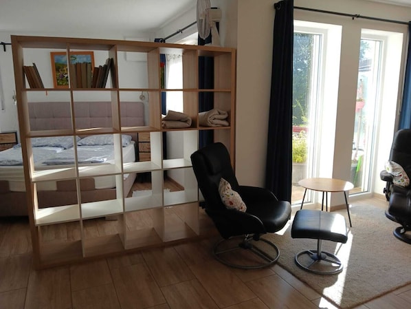 A room with a bed, a black chair, a small table, and a large window with curtains.