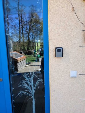 Une porte dotĂ©e dâun panneau de verre qui reflĂšte un jardin, un petit arbre et un interphone fixĂ© au mur.