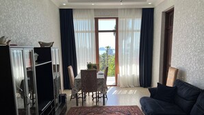 Una sala de estar con un sofá oscuro, una mesa de comedor con sillas, un mueble para la televisión y un ventanal con cortinas.
