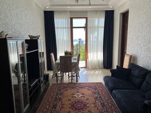 Un salon avec un canapé foncé, une table à manger avec des chaises, un meuble pour la télévision et une grande fenêtre avec des rideaux.