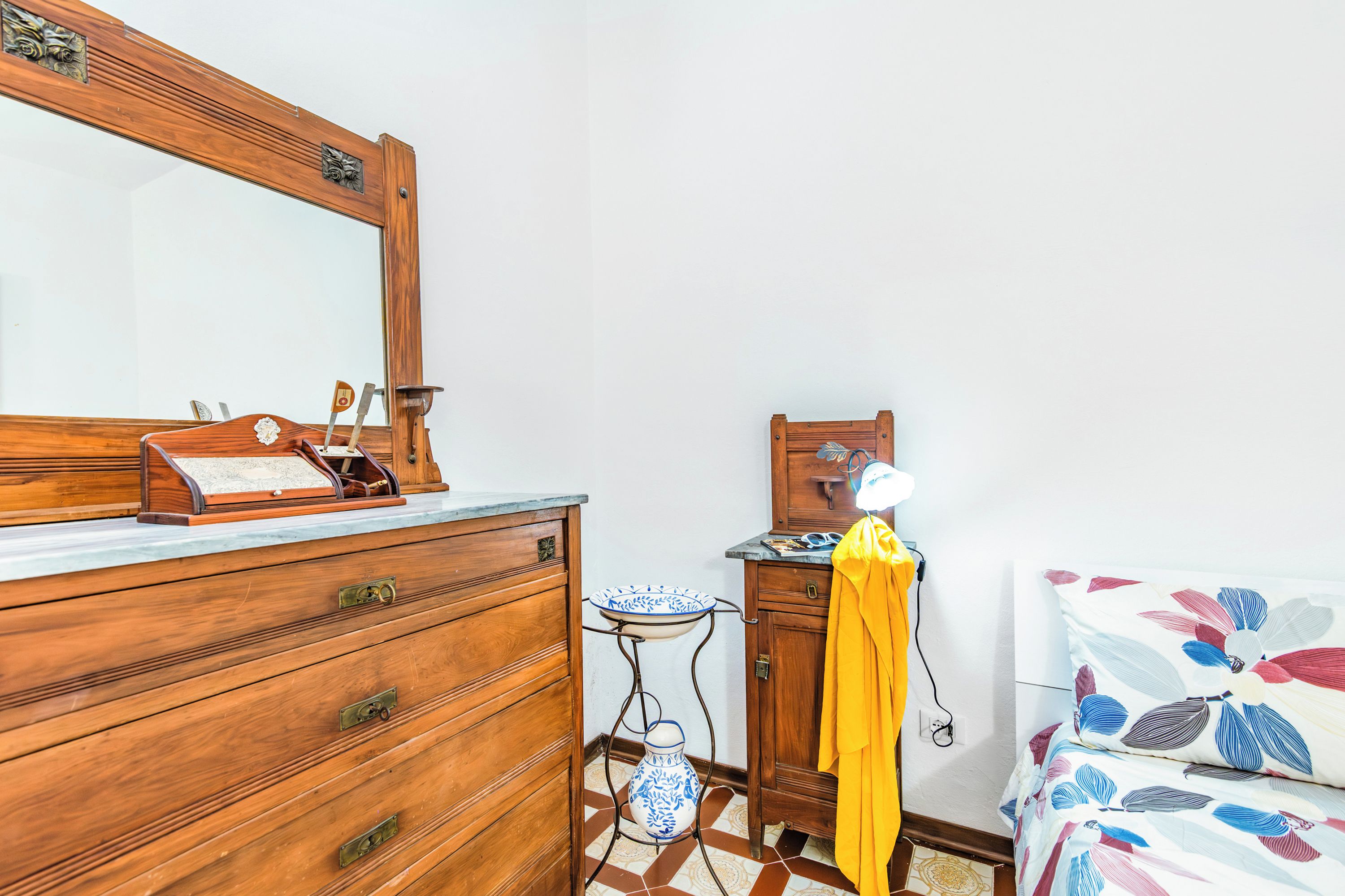 A wooden dresser with drawers, a mirror, a small table with a blue and white vase, and a bed with a floral bedspread.