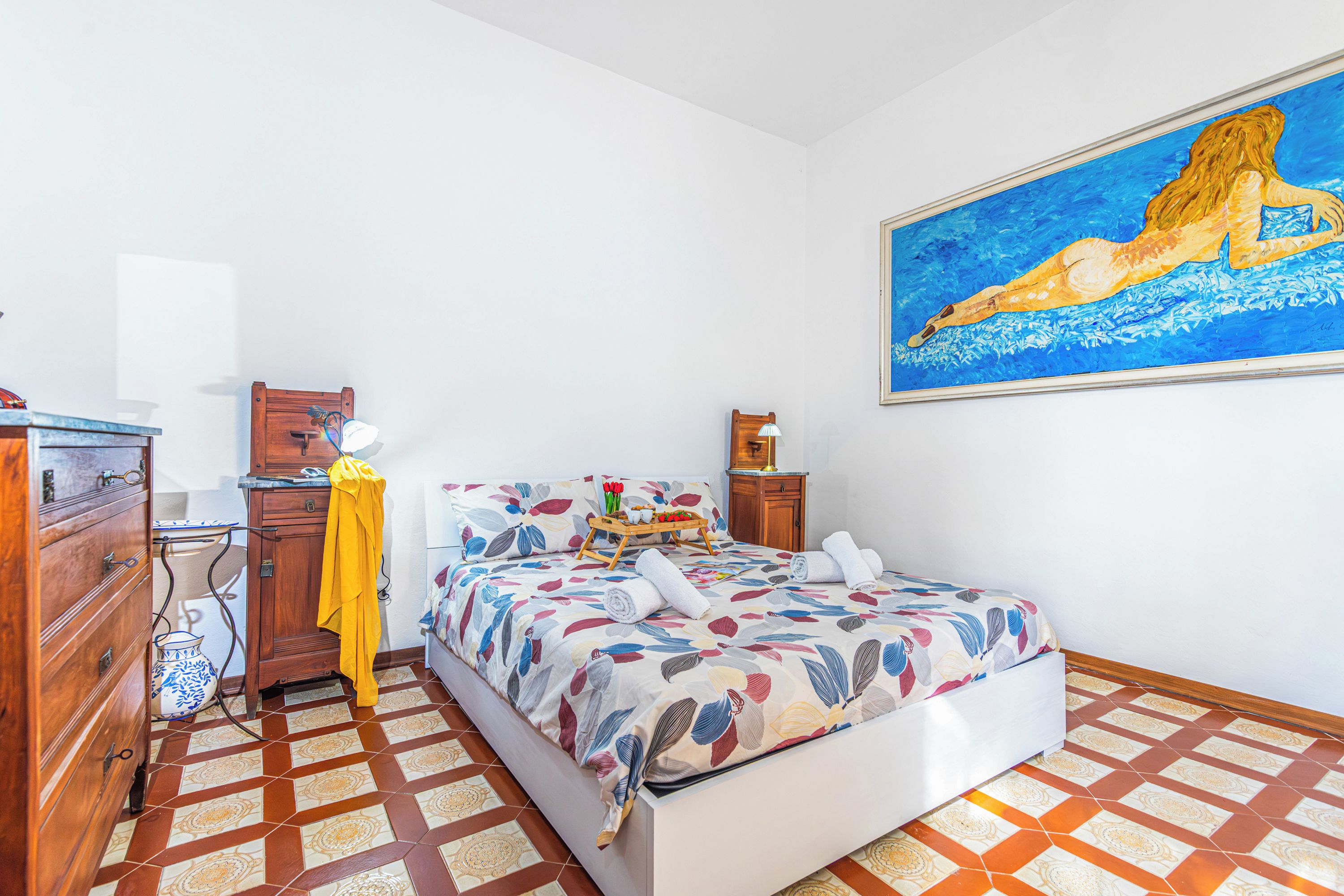 Un dormitorio con cama, cómoda de madera, mesita de noche, un cuadro y suelo de baldosas.
