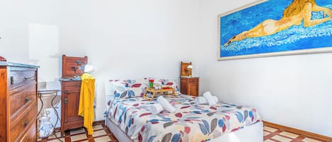 A bedroom with a bed, a wooden dresser, a nightstand, a painting, and a tiled floor.