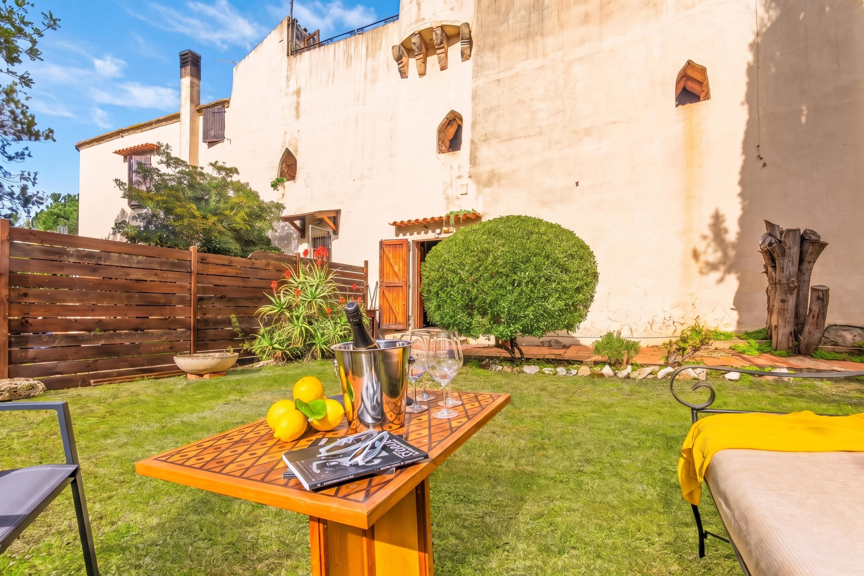A courtyard with a table set for a meal, a bottle of champagne, and a glass. There is a building with a white facade and a wooden fence in the background.