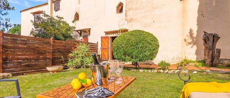 A courtyard with a table set for a meal, a bottle of champagne, and a glass. There is a building with a white facade and a wooden fence in the background.