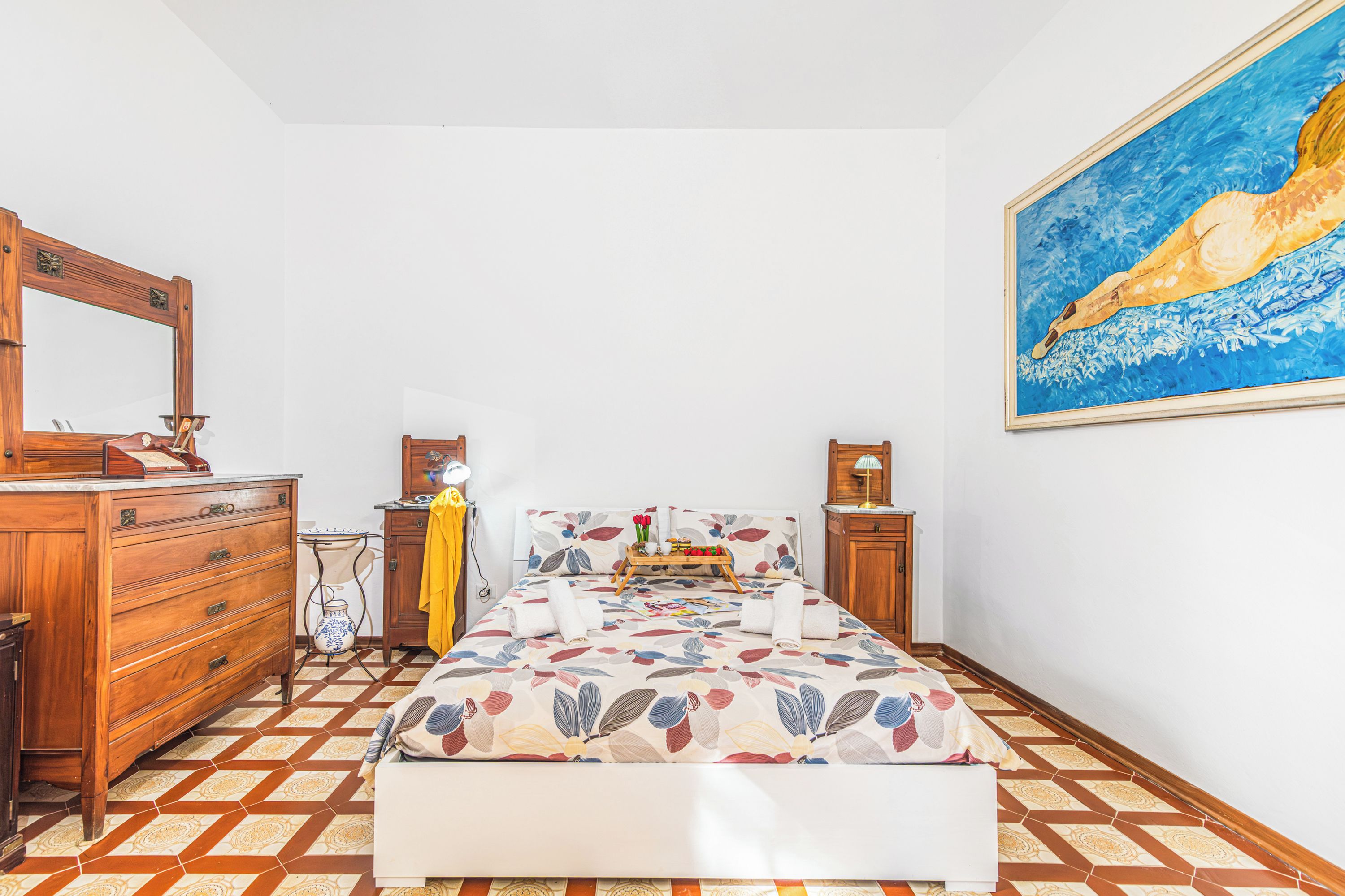 A bedroom with a bed, wooden dresser, bedside tables, a mirror, and a painting on the wall.