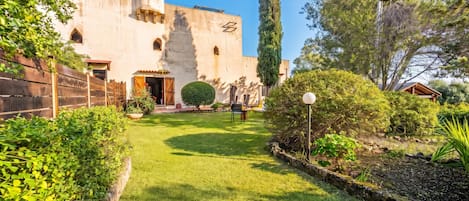 Un patio con un edificio que presenta una cúpula y ventanas de arco, rodeado de vegetación cuidada y un cielo despejado.