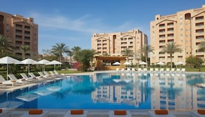 A swimming pool with lounge chairs, palm trees, and multi-story buildings in the background. - MISK Marina Valley Alamein (El Alamein)