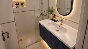 Een moderne badkamer met een glazen douchecabine, een witte wastafel met donker aanrecht, en een ronde spiegel met blauwe lijst.